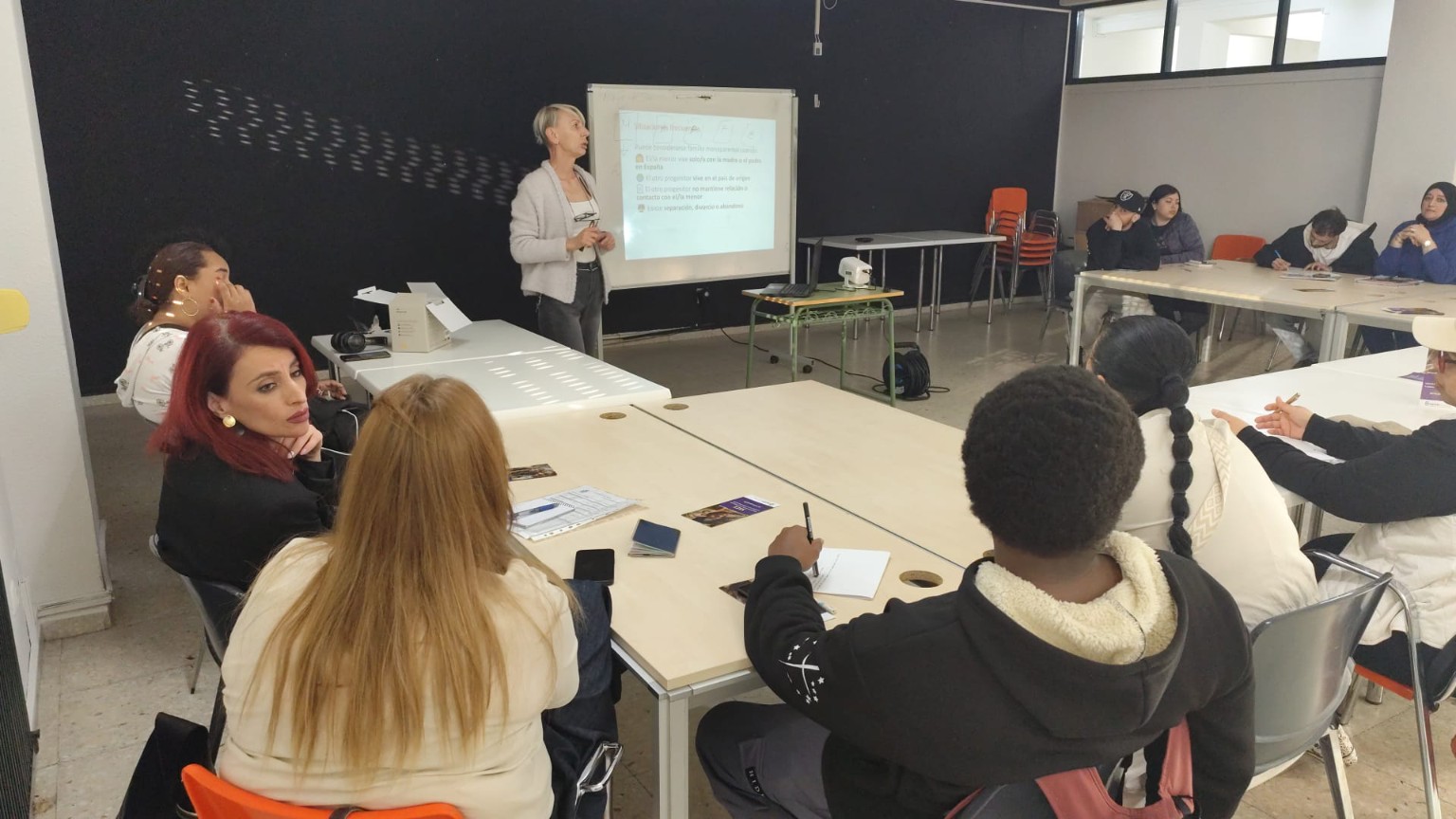 Una formadora explica una presentación ante un grupo de participantes sentados en un aula.
